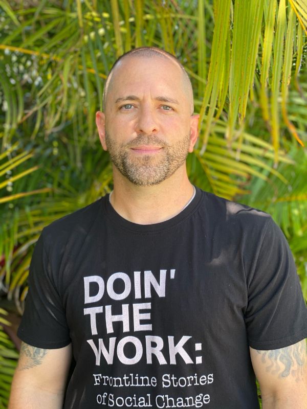 image of Shimon White Jewish male with short hair and beard in front of green palm trees in black Doin' The Work shirt